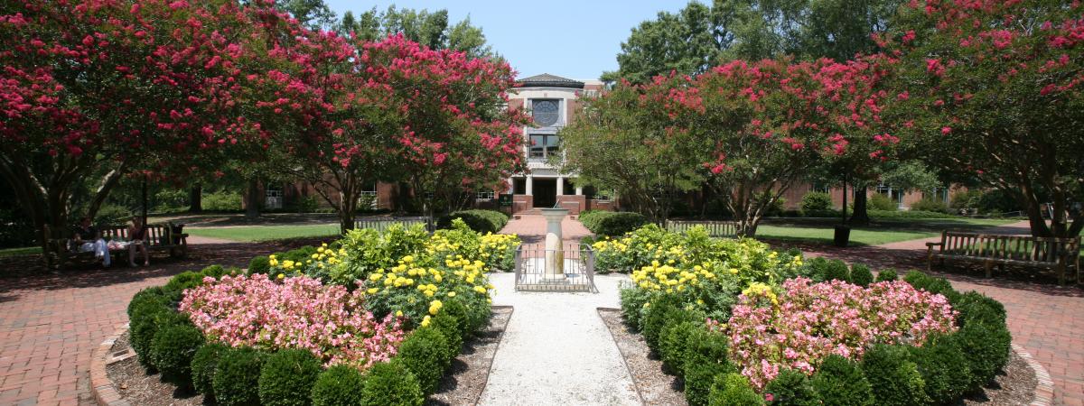 View of the sundial and circular bed of flowers, with the front entrance of the library in the distance framed by trees