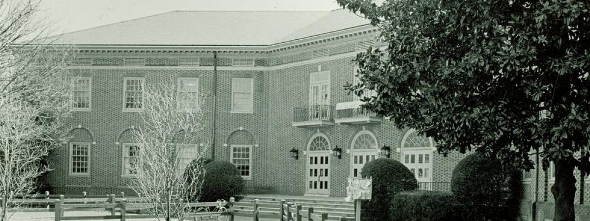 Black and white photo of two story, brick Campus Center with three arched doors and wood fenced sidewalk