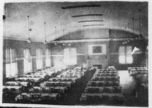 Black and white photo of the interior of Trinkle Hall with hanging overhead lights, large windows and long dining tables
