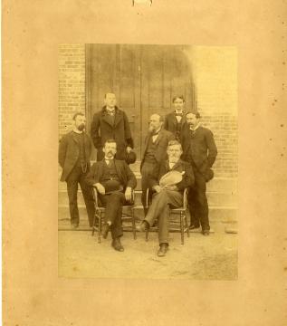 Yellowed, historic photograph of the Seven Wise Men, two seated and five standing wearing dark formal suits and posing in front of a wood door