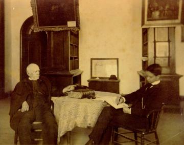 Ewell seated at a table talking to a younger person who is writing in a notebook