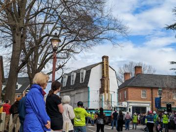 A white gambrel roof house loaded on a truck and being moved down a street with workers and members of the public looking on